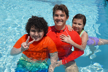 Three happy people, a young boy, a young man, and a young girl, smile in a swimming pool. The boy makes a peace sign, and the girl hugs the man in red.